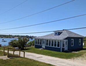 A small, single-story house with a gabled roof, a covered porch, and a gravel driveway leading to a wooden gate. - A working waterfront cottage in Downeast Maine, trails and beaches nearby (Jonesport)