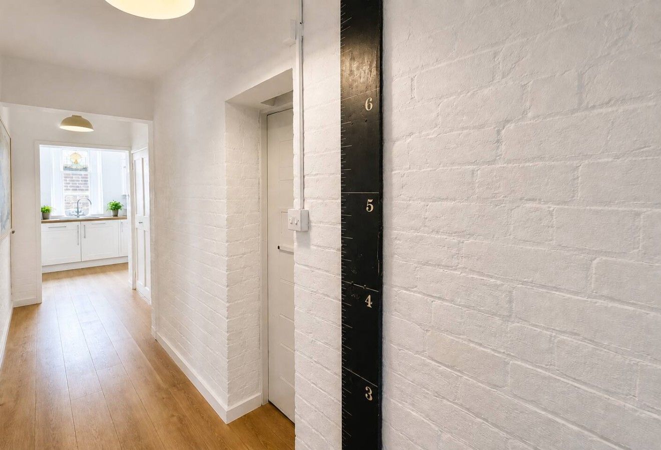 Un couloir aux murs en briques blanches, avec un plancher en bois et un instrument de mesure de la taille noir fixé au mur.