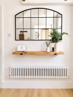 A white wall with a black-framed mirror, a wooden shelf with a framed certificate, a potted plant, and a white radiator.