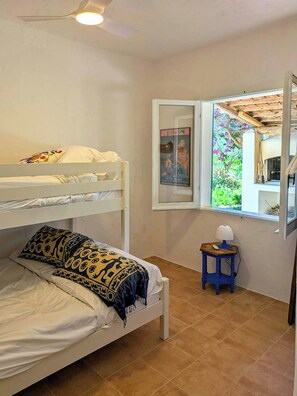 A room with a bunk bed, a small table with a lamp, a window with a view of greenery, and a framed picture on the wall.