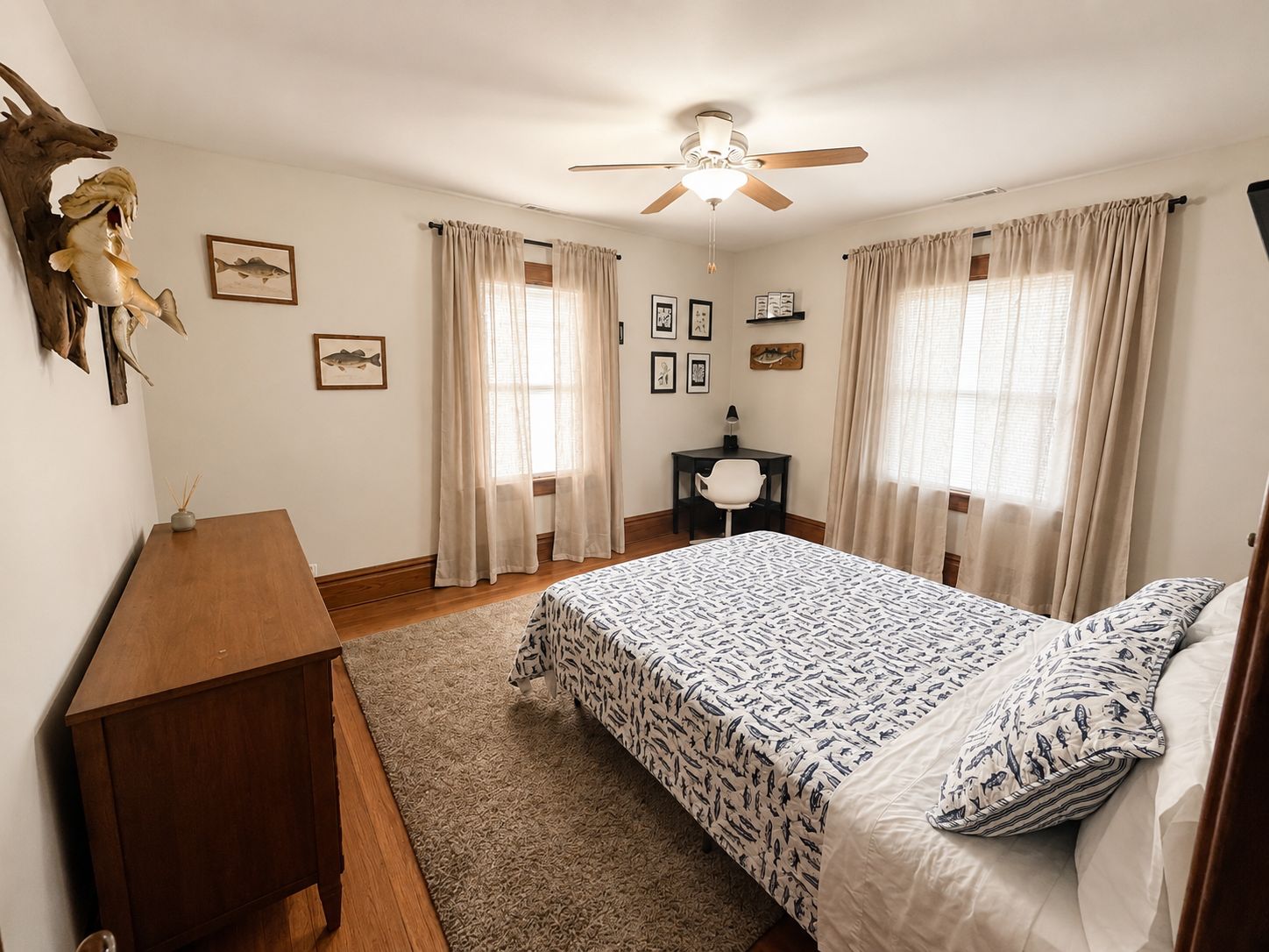 A bedroom with a bed, a desk, a chair, a ceiling fan, and two windows with curtains.