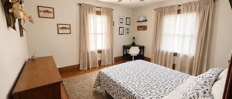 A bedroom with a bed, a desk, a chair, a ceiling fan, and two windows with curtains.