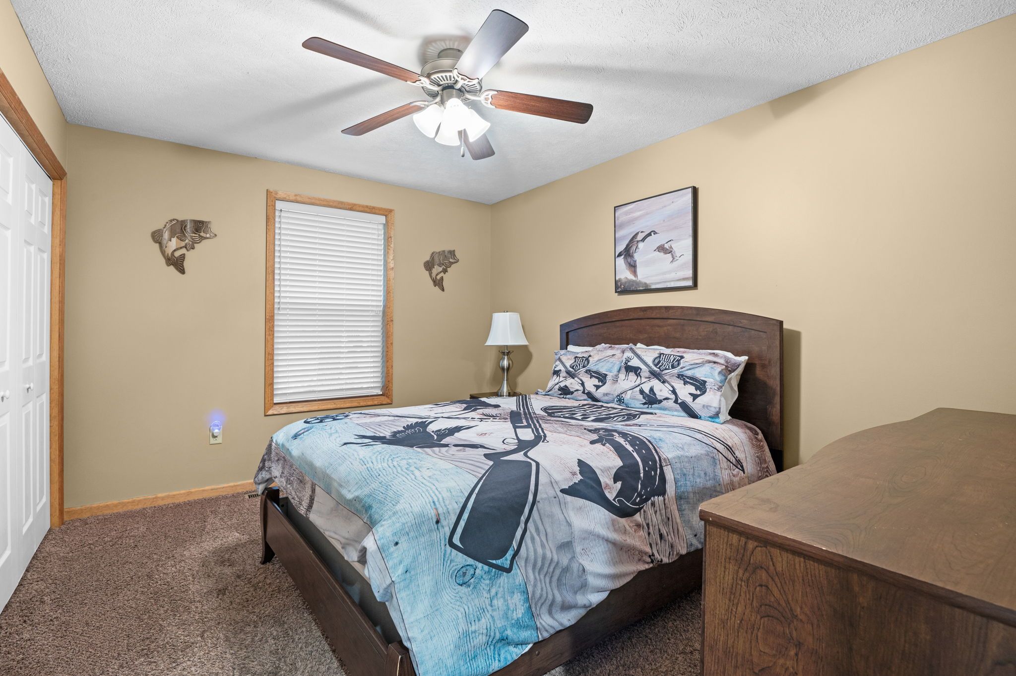 A bedroom with a bed, a nightstand, a ceiling fan, a window with blinds, and a wall-mounted picture.