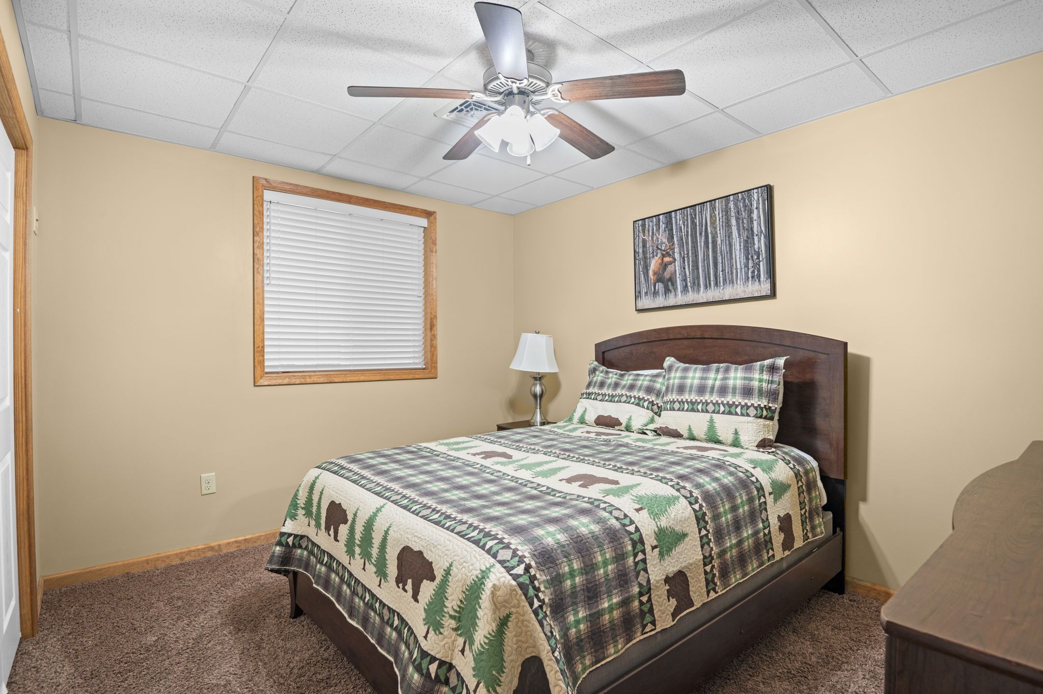 Een slaapkamer met een bed, een plafondventilator, een raam met zonwering, een bedlampje en een aan de muur bevestigd schilderij.