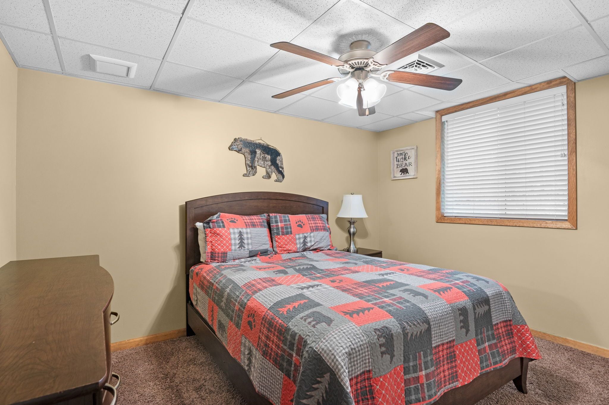 A bedroom with a bed, a ceiling fan, a wall-mounted lamp, a window with blinds, and a bear-themed wall decoration.