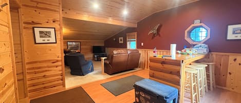 A cozy cabin interior with a wooden ceiling, a leather sofa, a small bar area, and a window with a view.