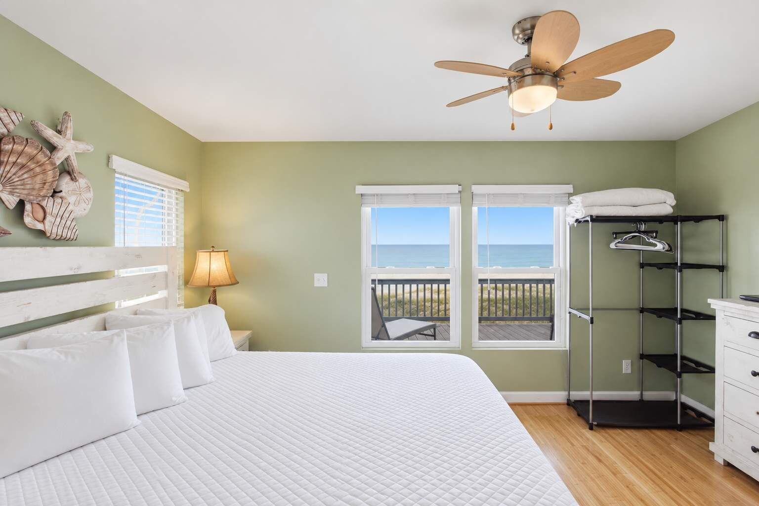 A bedroom with a bed, a ceiling fan, a nightstand, a lamp, a window with a view of the beach, and a storage unit.