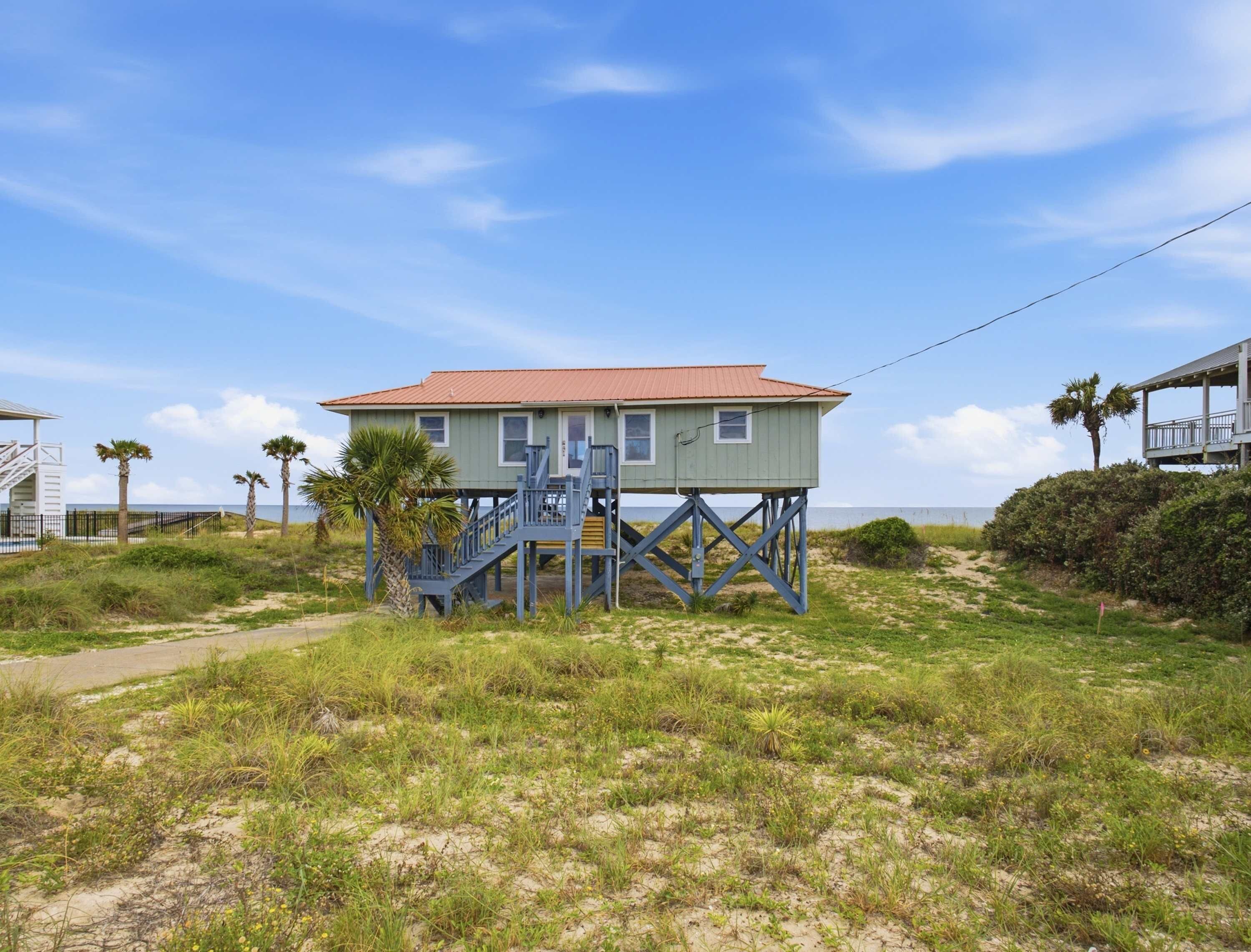 A beachfront house with a red roof, elevated on stilts, and a blue staircase leading to the entrance.