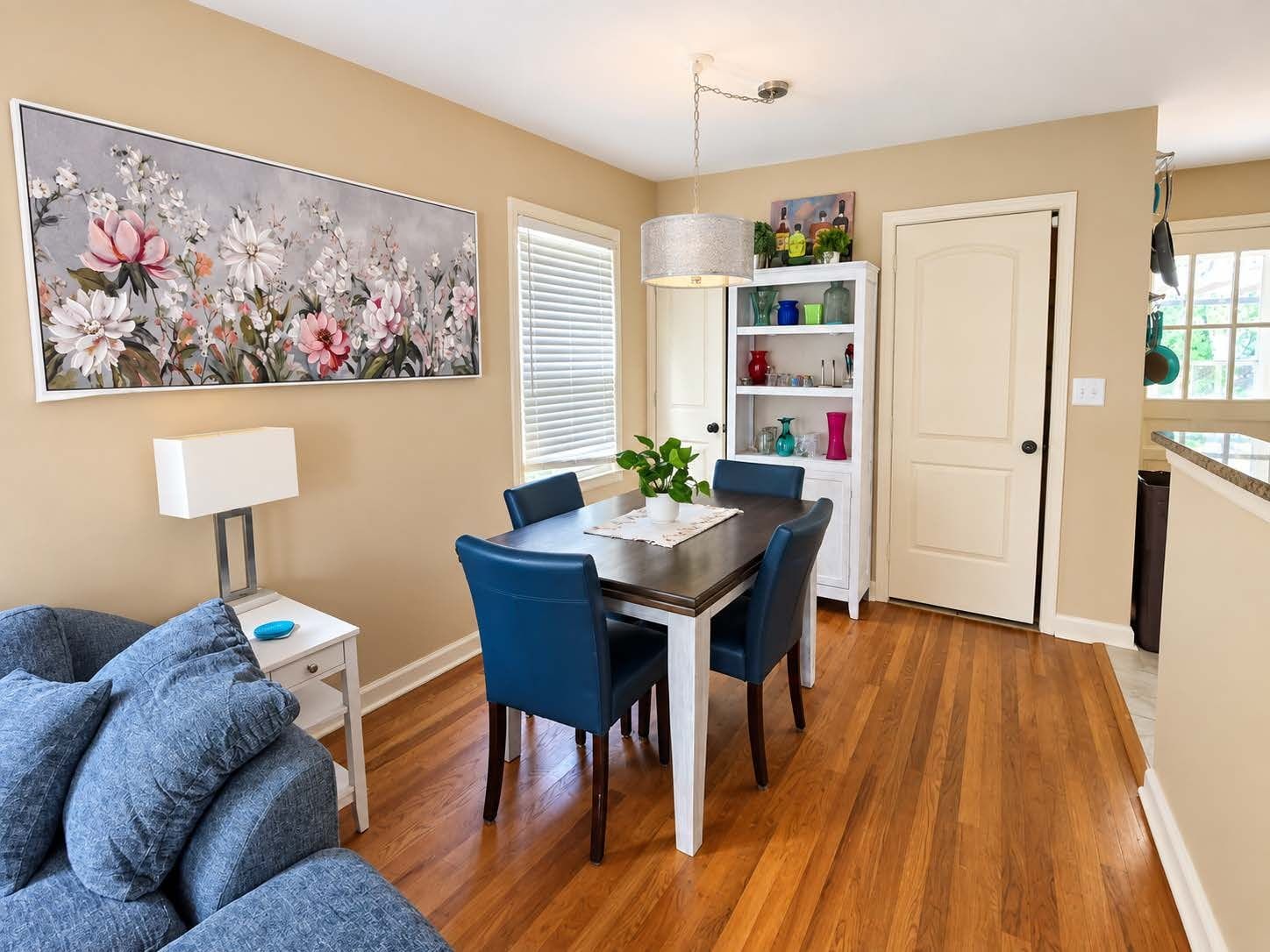 A dining area with a table, chairs, a lamp, and a floral painting on the wall.