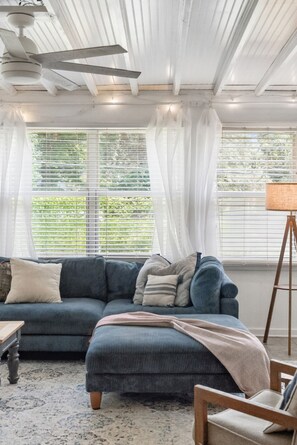 A room with a blue sofa, a wooden floor, a ceiling fan, and a window with blinds. - Renovated 4BR Home on James Island | Minutes to Do (Charleston)