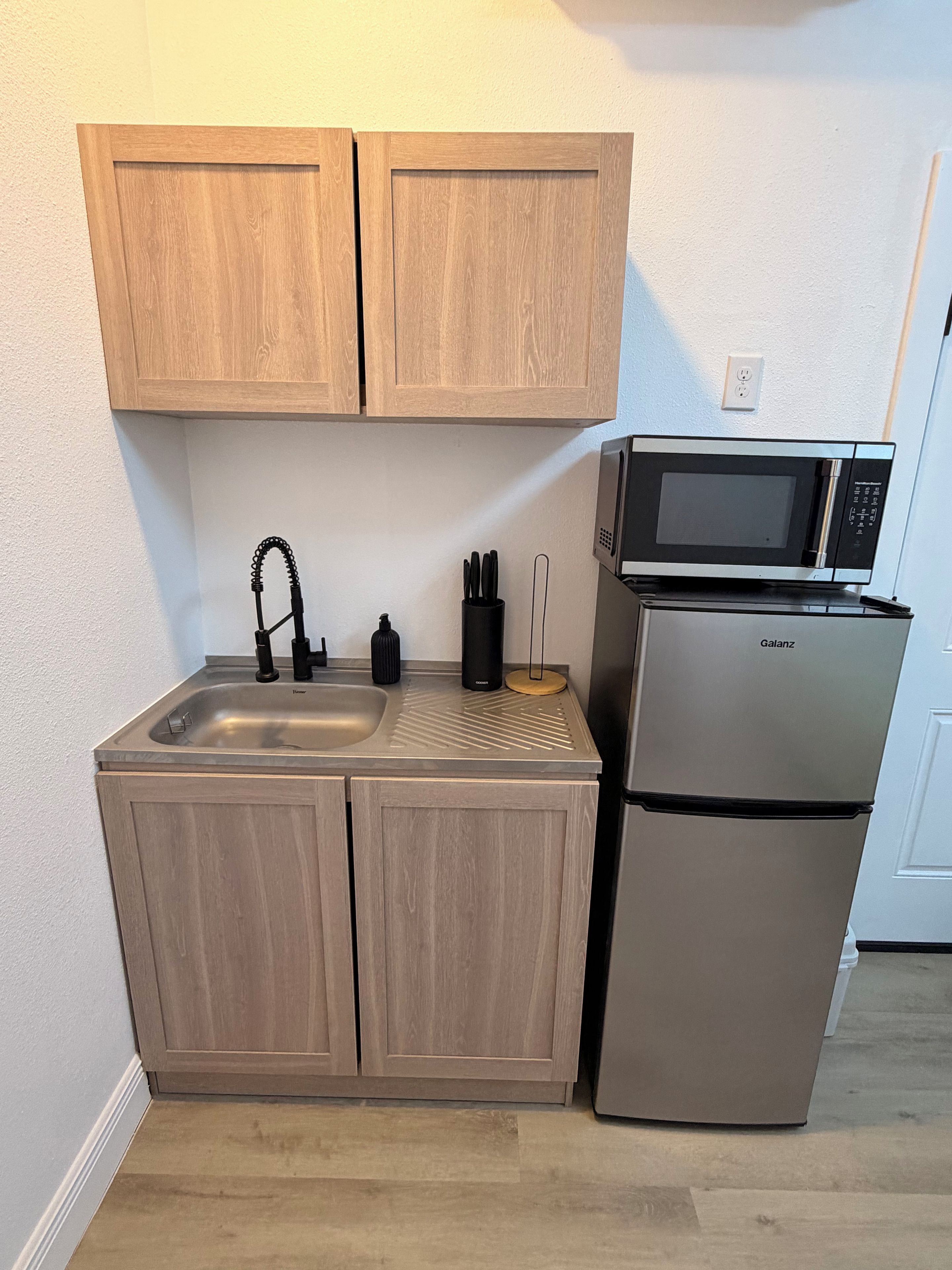 Eine kleine Küche mit einem Edelstahlkühlschrank, einer Mikrowelle, einem Spülbecken und Holzschränken.