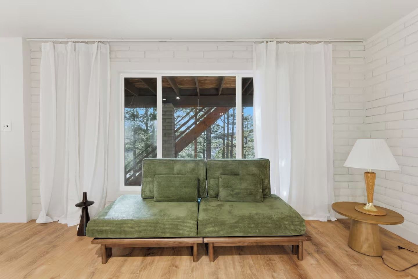 A room with a green sofa, a wooden side table with a lamp, and a view of trees through a window with white curtains.