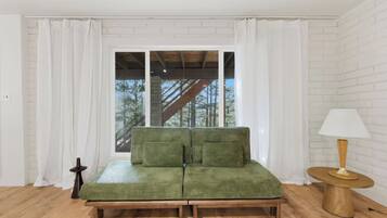A room with a green sofa, a wooden side table with a lamp, and a view of trees through a window with white curtains.