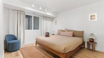 Un dormitorio con cama, una silla, una mesita de noche con lámpara y una ventana con cortinas.