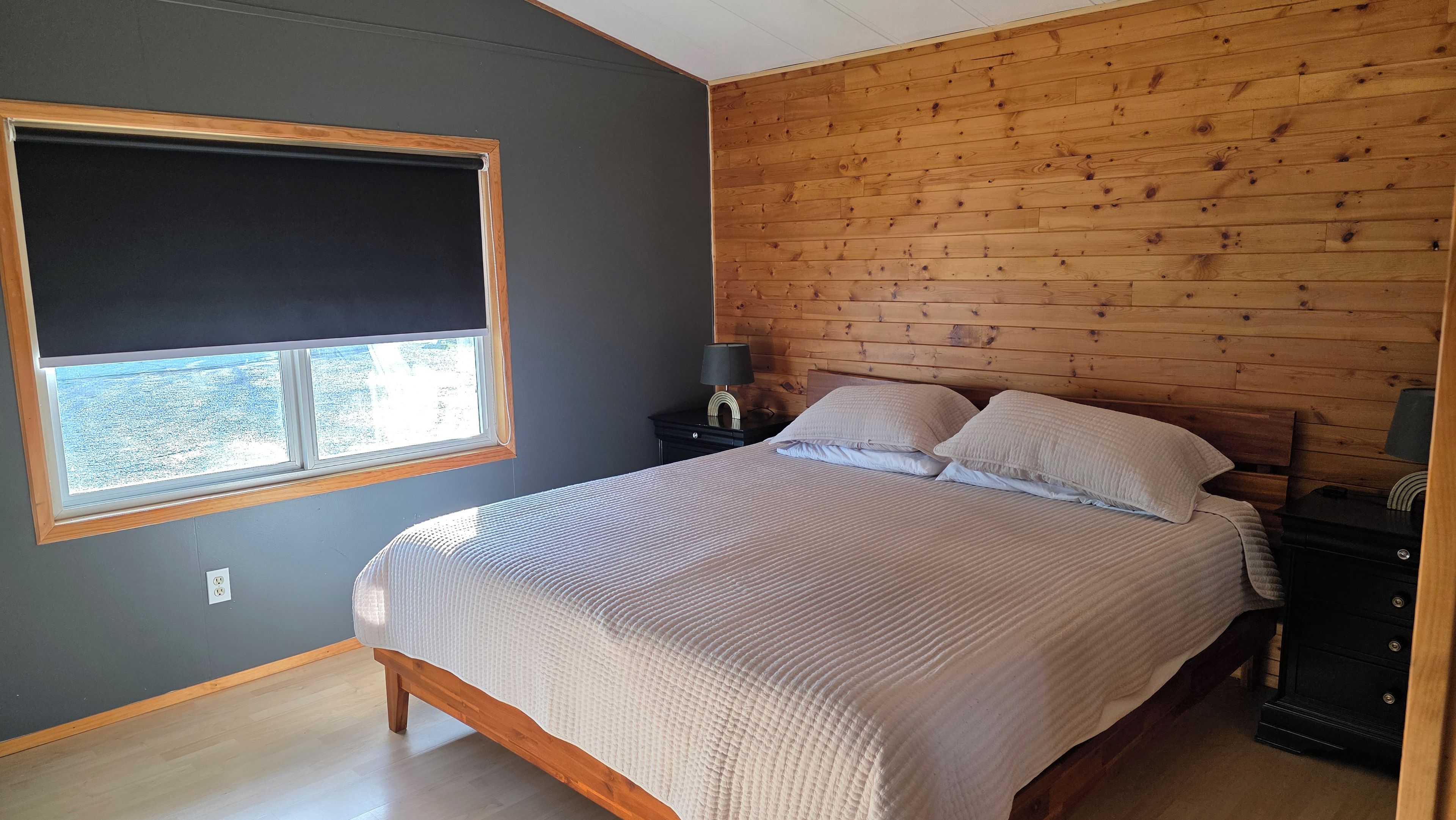A bedroom with a bed, a nightstand, a lamp, a window, and a wooden wall.