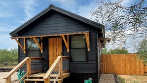 Une petite cabane de couleur sombre, dotée d’un escalier en bois menant à une porte d’entrée, est entourée d’une zone recouverte de gravier et agrémentée de deux cactus verts.