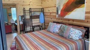 L’intérieur d’une cabane en bois, avec un lit, un portant à vêtements suspendu et un tableau accroché au mur.