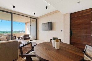 A modern living room with a round wooden table, a flat-screen TV mounted on the wall, and a large sliding glass door leading to an outdoor area. - Sáasil: Tranquil Oasis w/ Golf course views (Playa del Carmen)