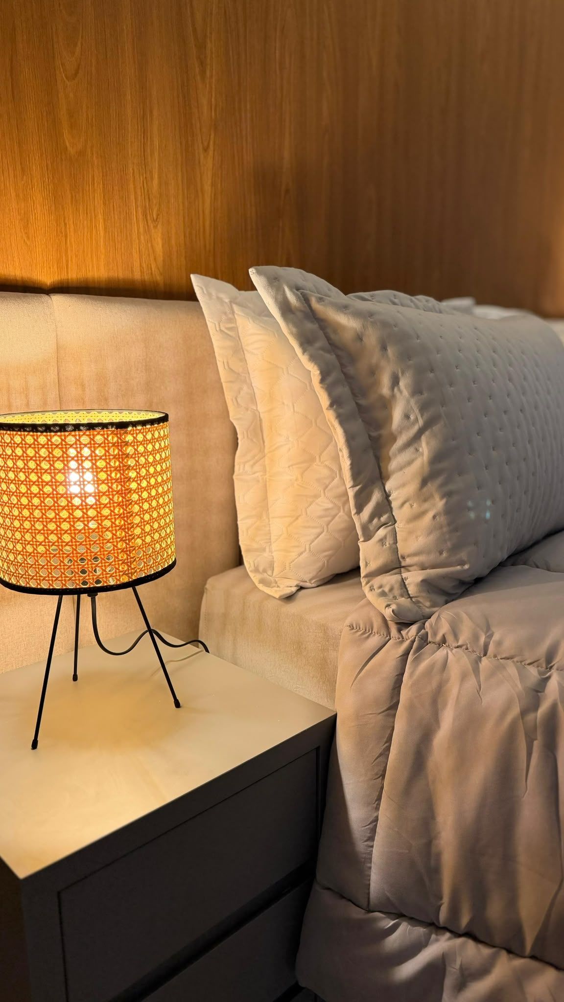 Una cama con colcha blanca y dos almohadas, una mesita de noche con lámpara y un cabecero de madera.