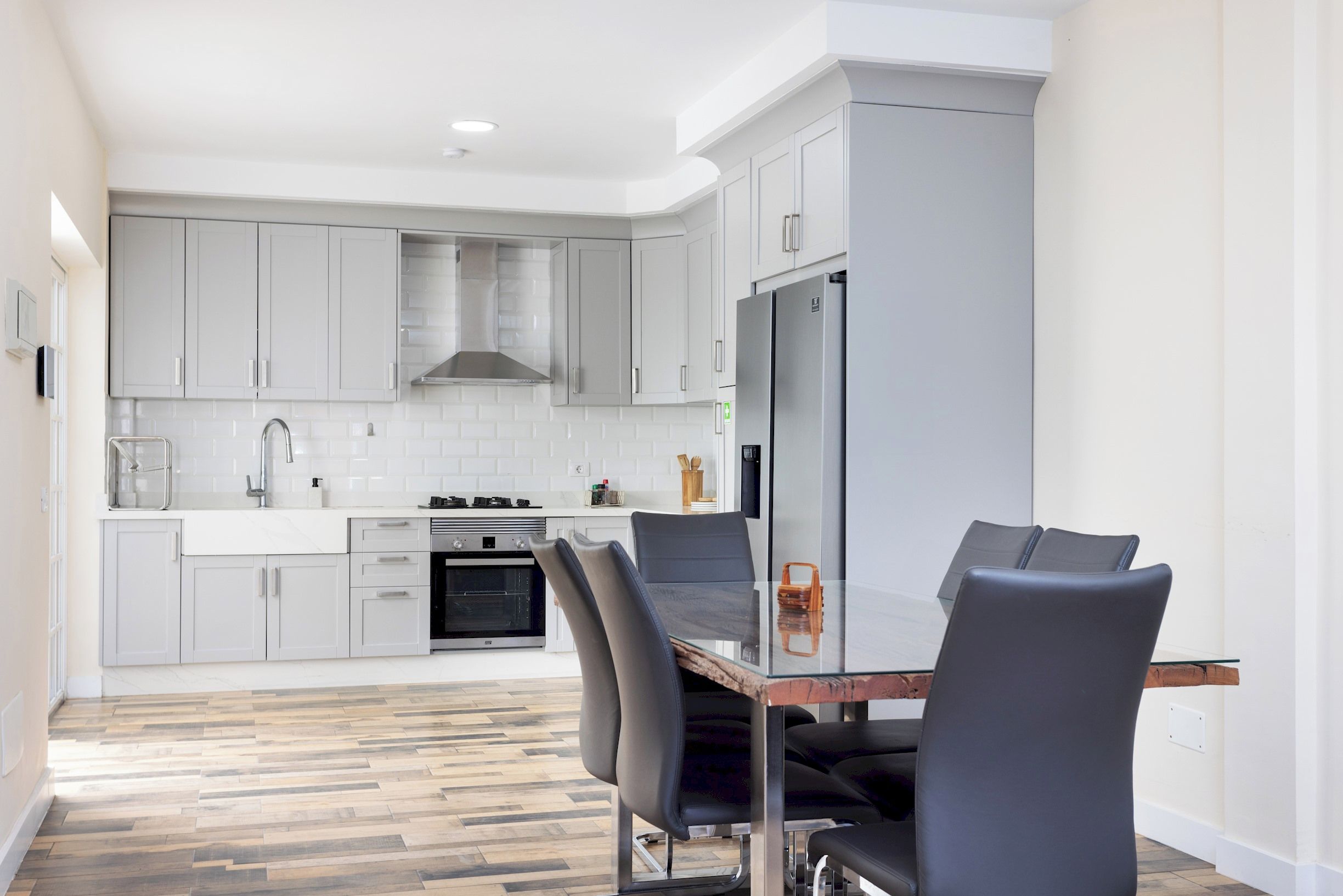 A modern kitchen with grey cabinets, a stainless steel refrigerator, a built-in oven, and a dining area with a glass table and black chairs.