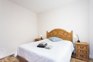 A neatly made bed with a white sheet and two folded towels, a wooden headboard with a decorative design, two bedside tables with lamps, and a wooden floor. - TenoHomes Spacious Buenavista House & BBQ (Buenavista del Norte)