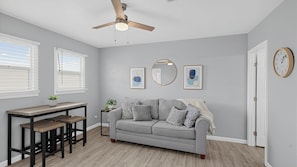 Une pièce avec un canapé gris, une table en bois avec des bancs, un ventilateur de plafond et deux fenêtres équipées de stores.