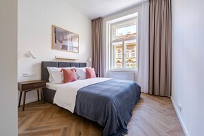 Ein Hotelzimmer mit Bett, Nachttisch, Wandlampe, einem Fenster mit Vorhängen und Blick auf Gebäude.