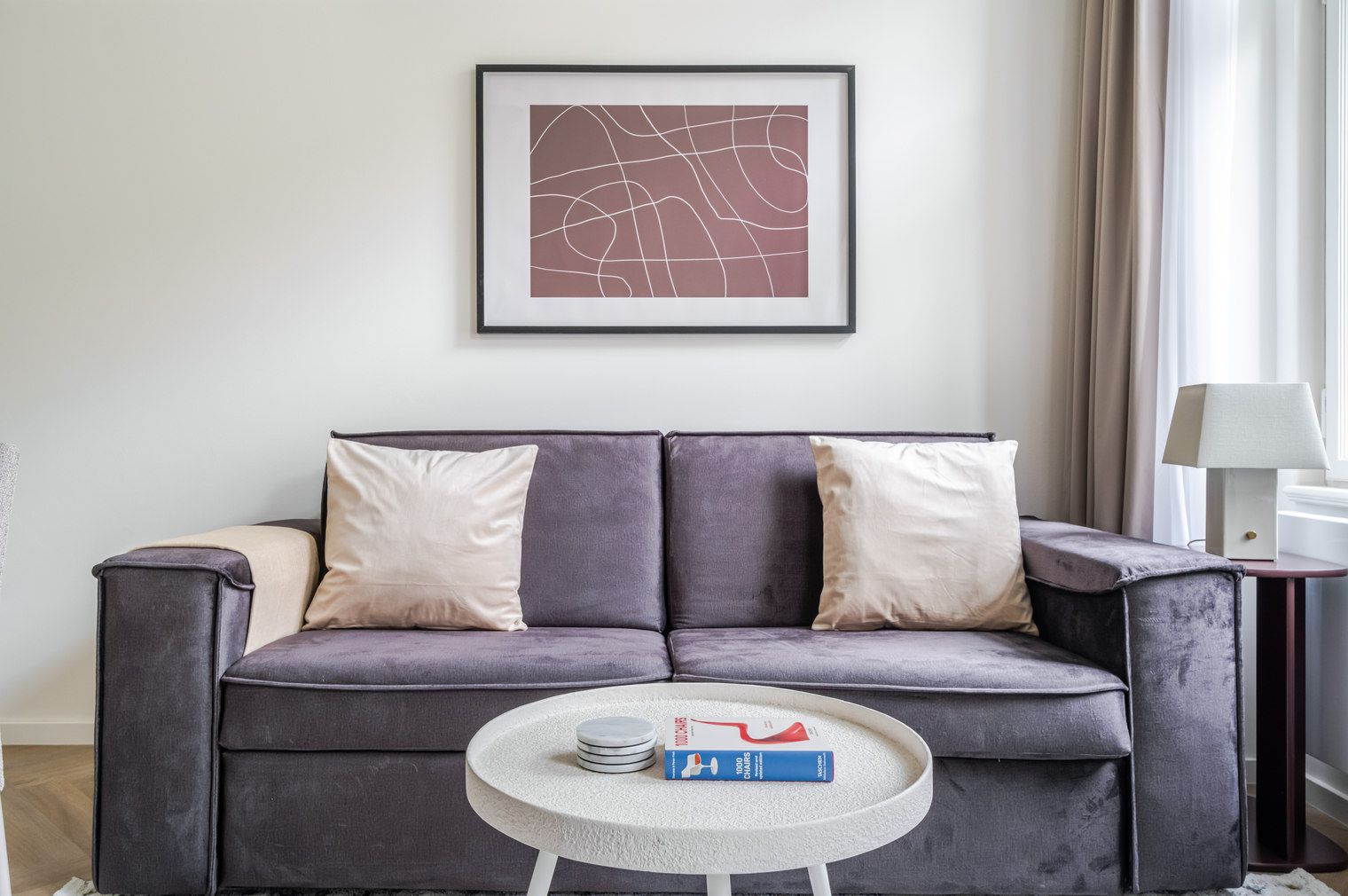 Un salon moderne avec un canapé gris, deux coussins blancs, une œuvre d’art abstraite encadrée, une petite table ronde sur laquelle se trouve un livre, et une lampe sur une table d’appoint.