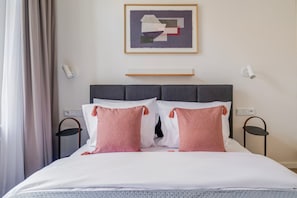 Ein ordentlich bezogenes Bett mit weißen und rosafarbenen Kissen, ein grauer Kopfteil und ein gerahmtes abstraktes Gemälde darüber.