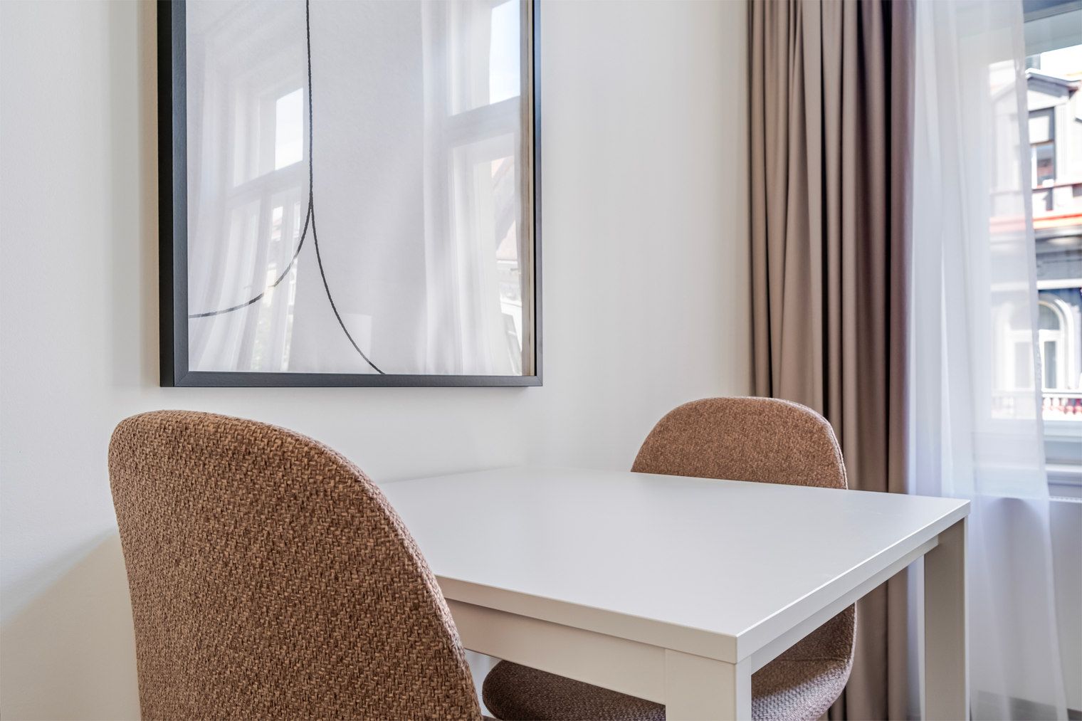 Una habitación con una mesa blanca y dos sillas marrones, una obra de arte abstracta enmarcada en la pared, y una ventana con cortinas.