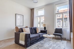 Ein modernes Wohnzimmer mit einer Couch, einem Sessel, einem kleinen Tisch und einem Fenster mit Vorhängen.