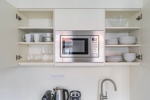 A kitchen with a built-in microwave, shelves with white cups and glasses, and a kettle. - Blueground | Vinohrady, view, nr Riegrovy sady (PRG-122) (Prague)