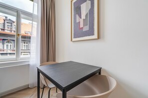 A room with a table, a chair, a window with curtains, and a framed picture on the wall. - Blueground | Vinohrady, view, nr Riegrovy sady (PRG-122) (Prague)