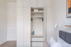 A bedroom with a bed, a grey headboard, a white wardrobe with open doors, and a wall-mounted light fixture. - Blueground | Vinohrady, view, nr Riegrovy sady (PRG-122) (Prague)