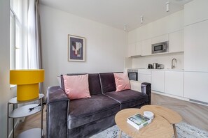 A modern living room with a sofa, a coffee table, a lamp, and a kitchen area with appliances. - Blueground | Vinohrady, view, nr Riegrovy sady (PRG-122) (Prague)