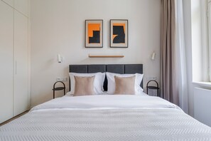 Una camera d'albergo con un letto, due opere d'arte appese al muro, un comodino, una sedia e una finestra con tende.