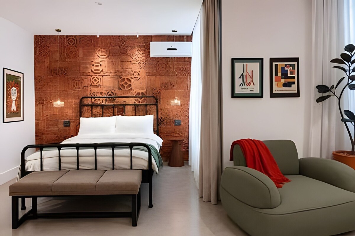 A modern bedroom with a brick-patterned headboard, a bed with white linens, a bench, a green sofa, and artwork on the walls.