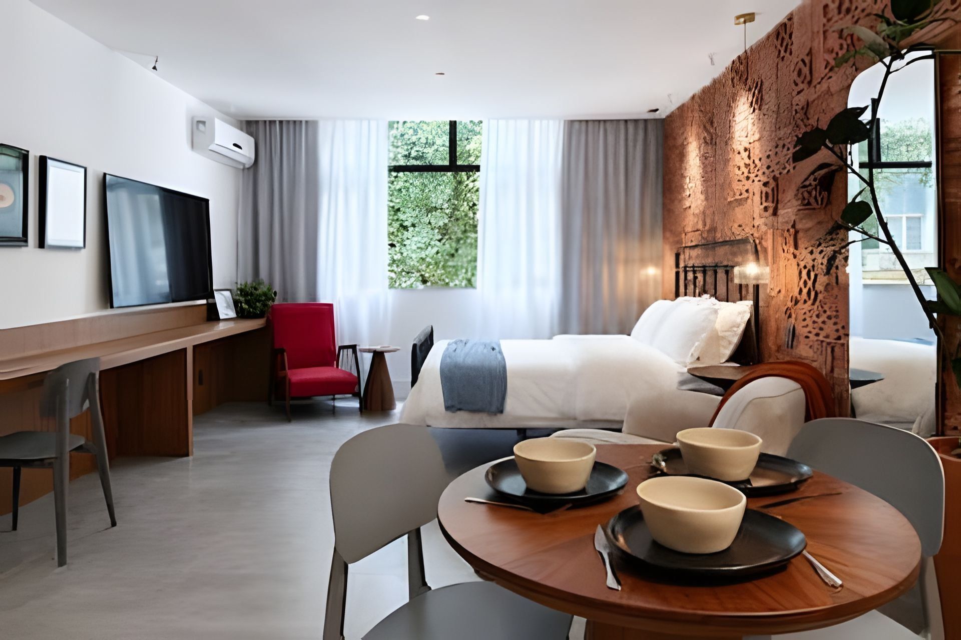 A modern hotel room with a bed, a desk, a red chair, and a dining table with bowls.
