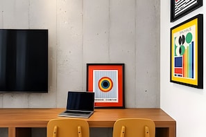 A modern office space with a wooden desk, a laptop, two yellow chairs, a framed abstract artwork, and a flat-screen TV mounted on a concrete wall. - Living Studios (Rio de Janeiro)