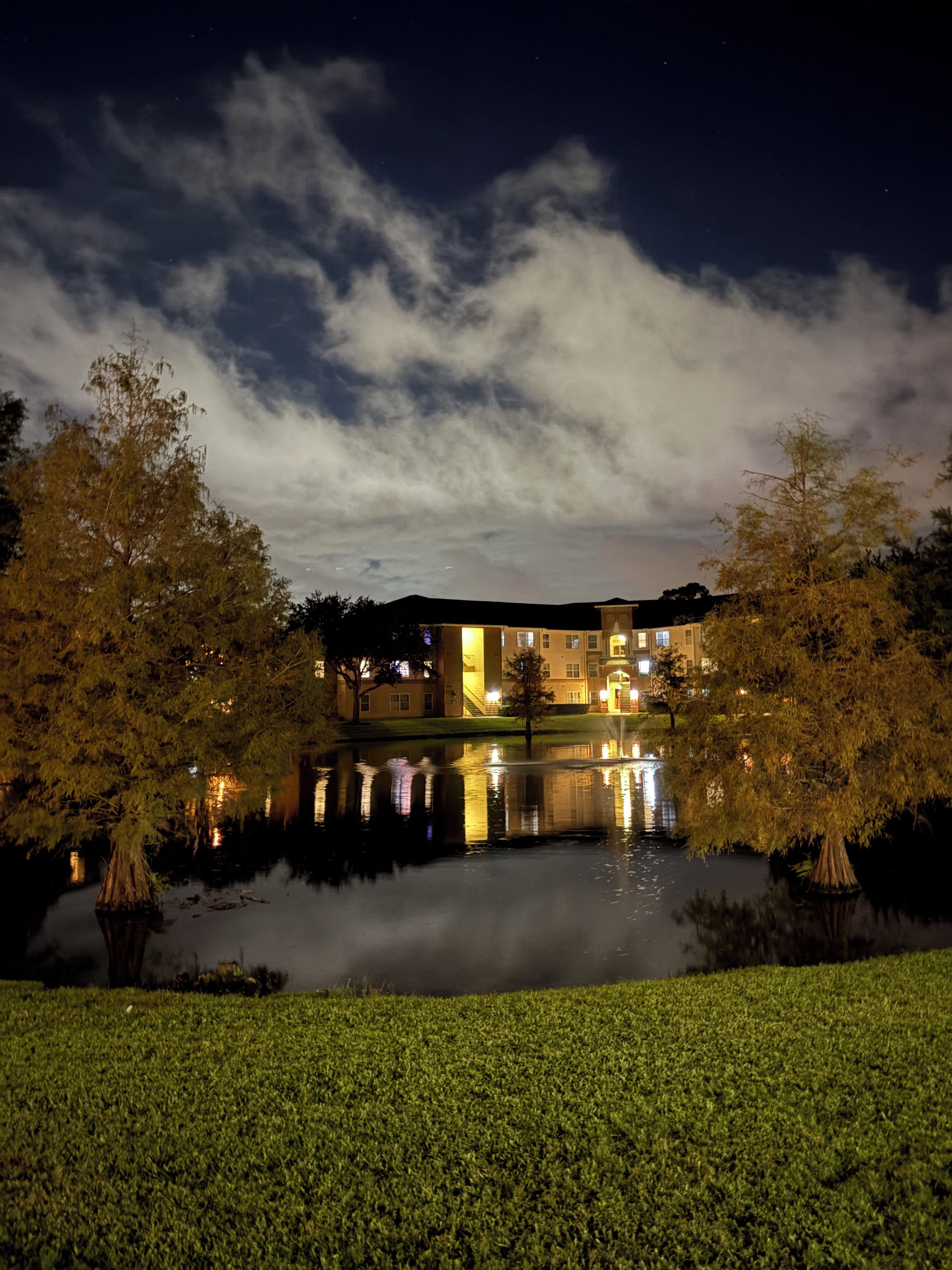 A building with multiple windows and a reflection in a calm body of water, surrounded by trees.