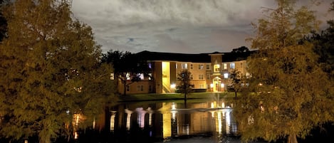 A building with multiple windows and a reflection in a calm body of water, surrounded by trees.