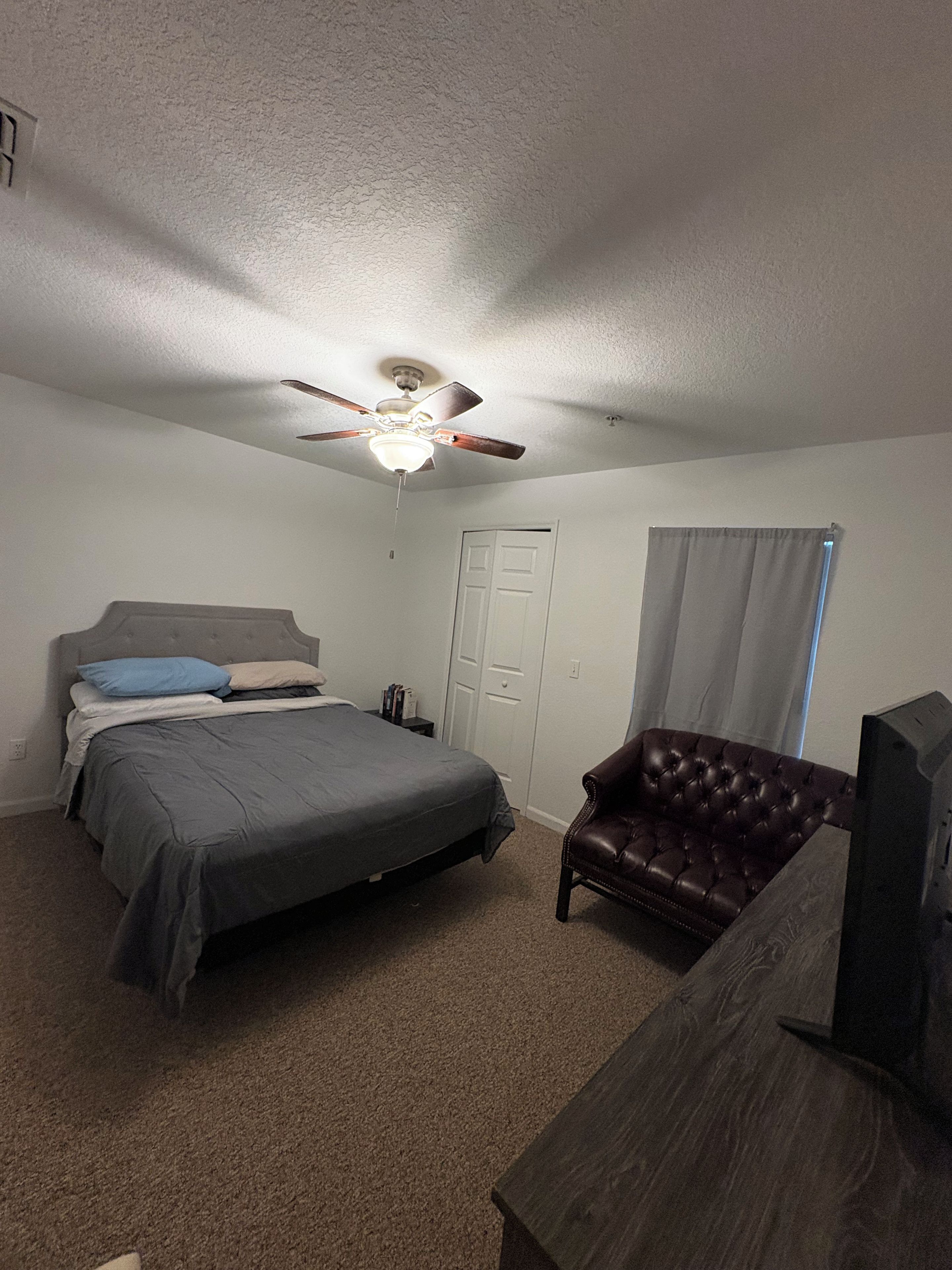 A bedroom with a bed, a ceiling fan, a dark brown leather chair, a wooden desk, and a television.