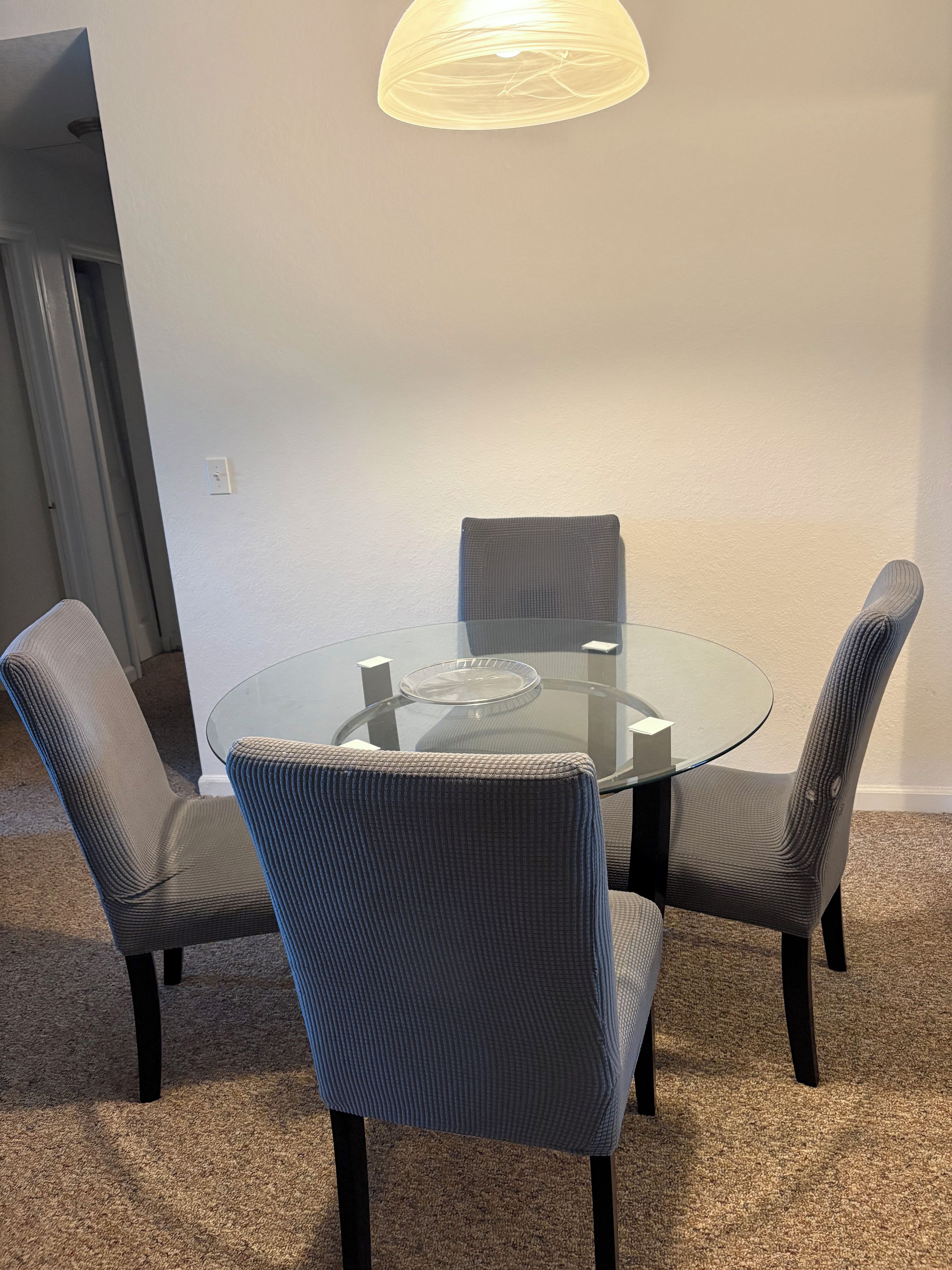 A round glass table with four chairs, a light fixture above, and a carpeted floor.