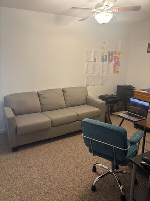 Une pièce avec un canapé beige, une chaise de bureau bleue, un bureau avec un ordinateur portable, une imprimante et un ventilateur de plafond.