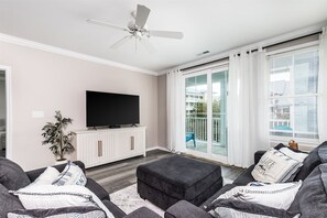 A modern living room with a flat-screen TV, a ceiling fan, and a sliding glass door leading to a balcony. - Sunset Island 37 Fountain Drive West, Unit 3E (Ocean City)
