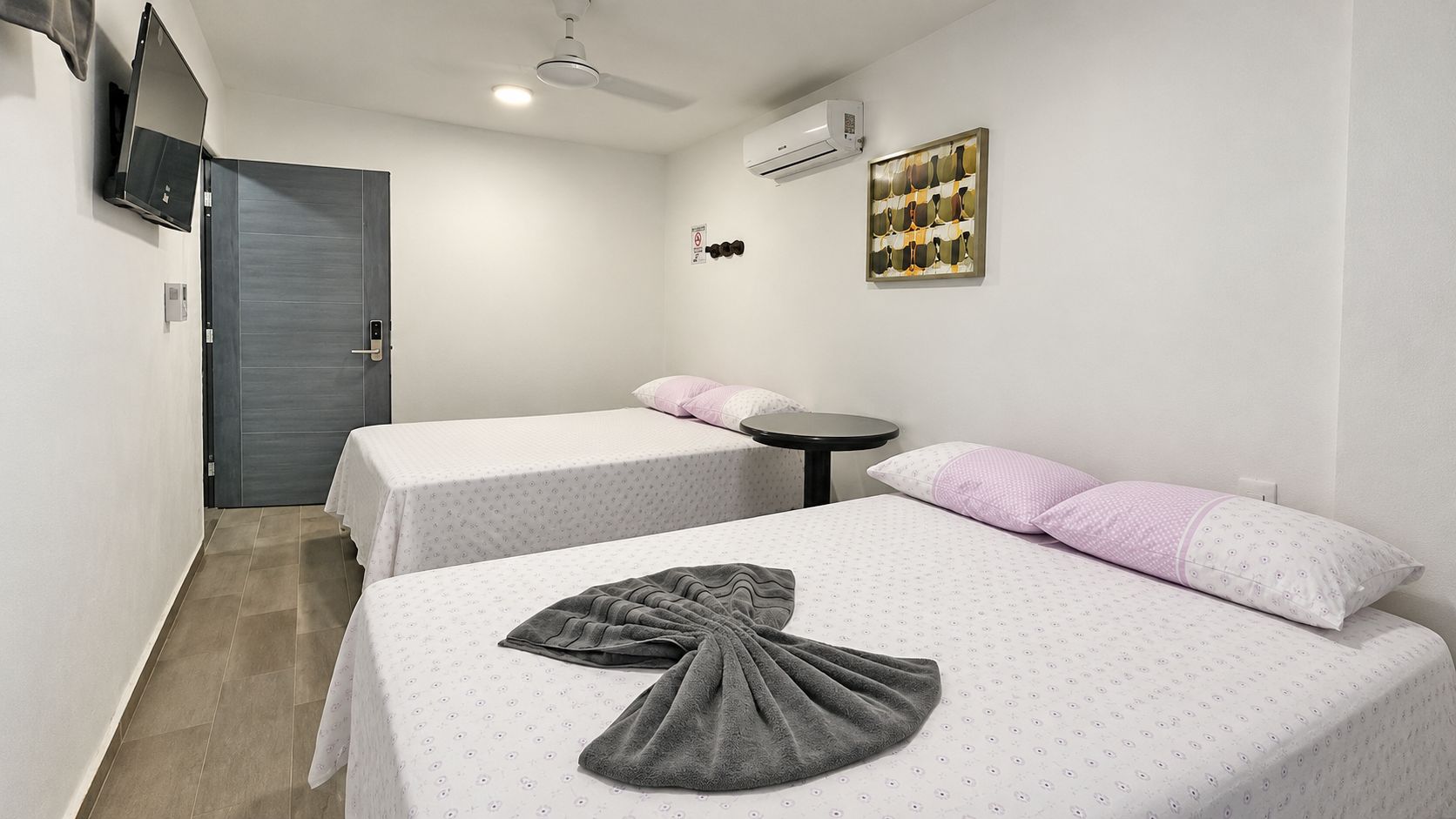 A hotel room with two beds, a small table, an air conditioning unit, and a wall-mounted television.