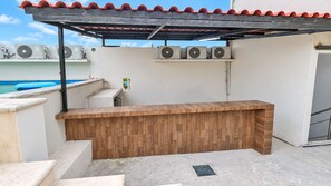 An outdoor bar area with a wooden counter, air conditioning units, and a pool in the background.