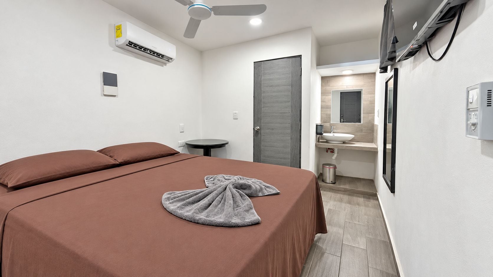 Ein Hotelzimmer mit Bett, Nachttisch, Wandklimaanlage, Deckenventilator und Badezimmer mit Waschbecken und Spiegel.