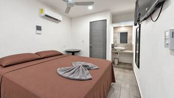 Ein Hotelzimmer mit Bett, Nachttisch, Wandklimaanlage, Deckenventilator und Badezimmer mit Waschbecken und Spiegel.