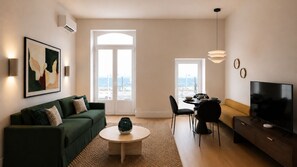 A modern living room with a green sofa, a round coffee table, a dining area with a table and chairs, a TV, and a large window with a view. - Ando Living - Tejo Palace - 101 (Lisboa)
